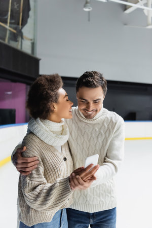 Smiling interracial couple hugging and using smartphone on ice rinkの写真素材