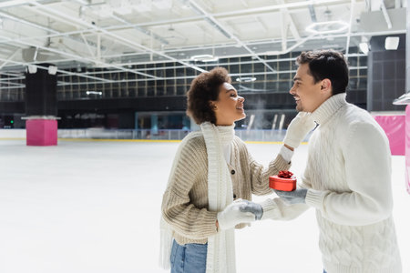 Side view of smiling man holding heart shaped gift box and hand of african american girlfriend on ice rinkの写真素材