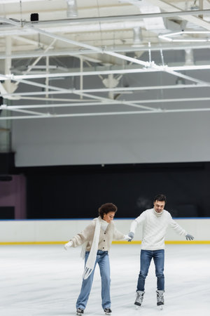Smiling multiethnic couple in gloves and sweaters ice skating on rinkの写真素材