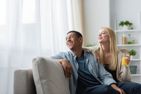 blonde woman with orange juice and smiling african american man sitting on sofa in kitchen and looking awayの写真素材