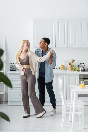 full length of joyful interracial couple smiling at each other while dancing in modern kitchenの写真素材