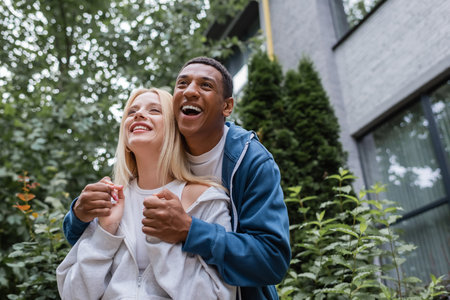 excited african american man holding hands of blonde girlfriend and laughing on streetの写真素材