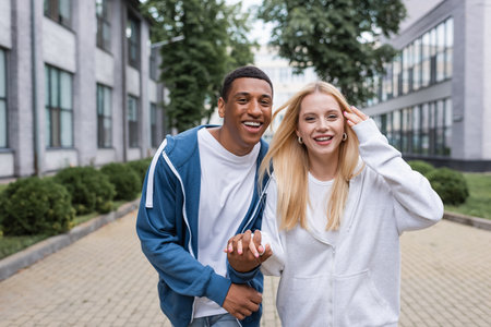 happy multiethnic couple in hoodies holding hands and looking at camera on urban street