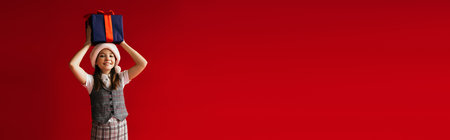 joyful girl in santa hat holding blue gift box above head isolated on red, bannerの写真素材