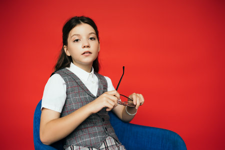 preteen girl in plaid vest sitting in armchair and holding eyeglasses isolated on redの写真素材
