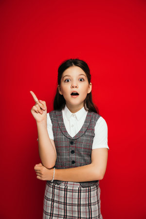 astonished brunette girl in plaid vest looking at camera and pointing with finger isolated on red, bannerの写真素材