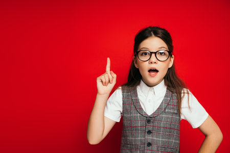 astonished girl in eyeglasses showing idea gesture and looking at camera isolated on red
