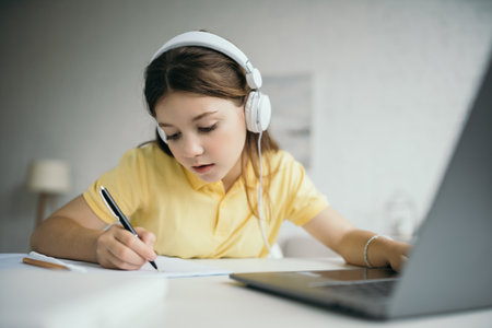 preteen schoolgirl in wired headphones writing in notebook near computer on blurred foreground