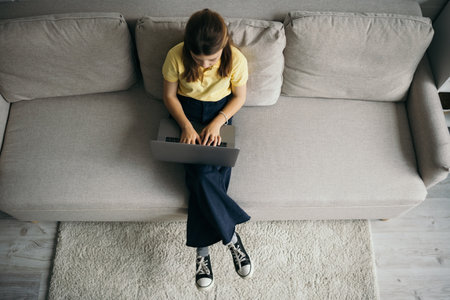 top view of preteen girl using laptop on modern sofa at homeの写真素材