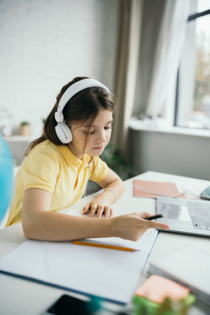 schoolgirl in headphones pointing with finger while studying near blurred laptop at home