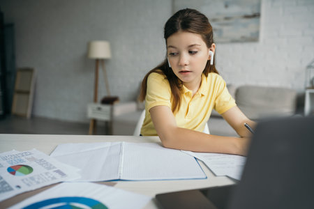 brunette girl in wireless earphones learning near notebook and computer on blurred foreground