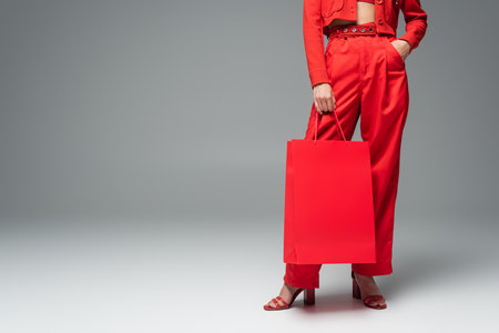 cropped view of woman holding shopping bag while standing with hand in pocket of red pants on grey backgroundの写真素材
