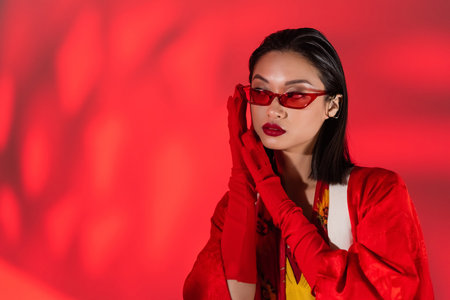 trendy asian woman in red gloves and trendy sunglasses holding hands near face on abstract red backgroundの写真素材