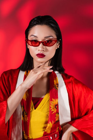 asian woman in trendy sunglasses and kimono cape with floral print scarf touching chin on abstract red backgroundの写真素材
