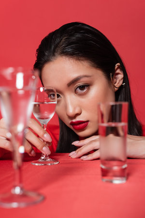 portrait of asian woman with makeup and ear cuff near glasses of clear water on blurred foreground isolated on redの写真素材