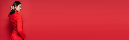 side view of brunette asian woman in trendy outfit touching hair on red background, banner