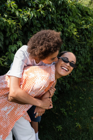 happy african american lesbian woman embracing cheerful girlfriend in stylish sunglasses laughing in...
