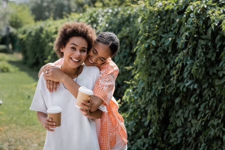 happy african american lesbian couple holding paper cups with coffee to go while hugging in parkの写真素材
