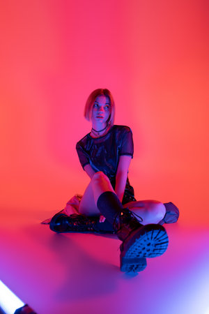 young woman in stylish leather boots looking away while sitting in neon light on purple and coral...