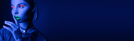 young woman with bright neon visage touching green lips and showing hush gesture on dark blue background, bannerの写真素材