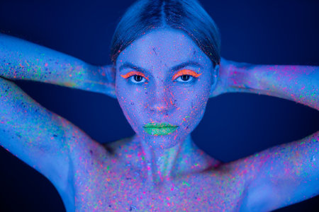 portrait of woman with vibrant neon makeup and bright paint splashes on body holding hands behind head isolated on dark blueの写真素材