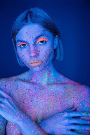 nude woman with neon makeup and bright paint on body covering bust with hands isolated on dark blueの写真素材