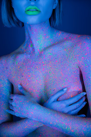 cropped view of nude woman with green neon lips and bright paint splashes on body covering breast isolated on dark blueの写真素材
