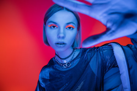 stylish woman in blue neon light posing with blurred outstretched hand on coral and pink background