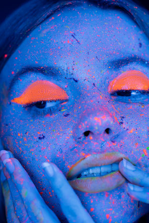 close up portrait of woman with fluorescent makeup touching face in blue neon lightの写真素材