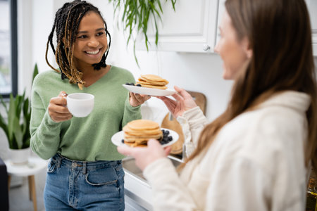 young woman holding plates with tasty pancakes near happy african american girlfriend with cup