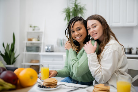 cheerful lesbian woman hugging smiling african american girlfriend during breakfastの写真素材