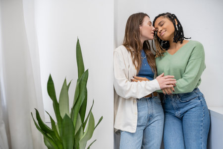 happy interracial and lesbian women smiling while hugging near green plant at home