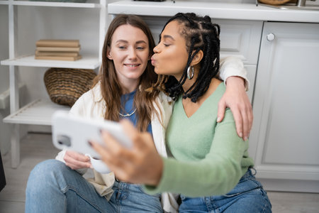 lesbian african american woman taking selfie while kissing cheek of girlfriend in kitchenの写真素材