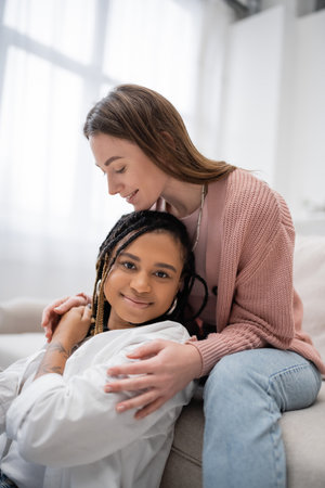 happy and lesbian woman sitting on couch and hugging tattooed african american girlfriend