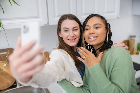 lesbian woman taking selfie while cheerful african american girlfriend showing engagement ring on fingerの写真素材