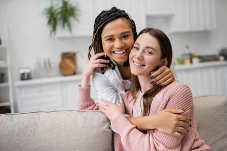 positive african american lesbian woman hugging joyful girlfriend in living roomの写真素材