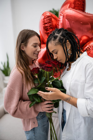 happy lesbian woman holding red roses near tattooed african american girlfriend and balloons on valentines dayの写真素材