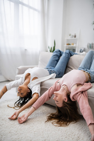 positive and multiethnic lesbian couple lying upside down on couch in living roomの写真素材