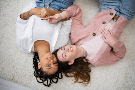top view of joyful and multiethnic lesbian couple lying on soft carpetの写真素材