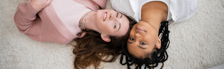 top view of positive and multiethnic lesbian couple lying on soft carpet, bannerの写真素材