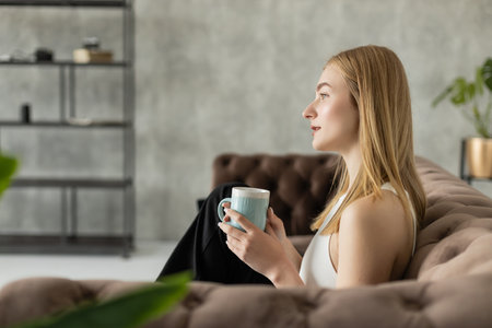 Side view of blonde woman holding cup of coffee and looking away on modern couch at homeの写真素材