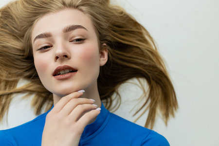 top view of blonde woman touching chin and looking away while lying on grey background