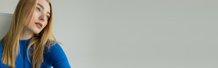 young and dreamy woman in blue zipped turtleneck looking away on grey background, bannerの写真素材