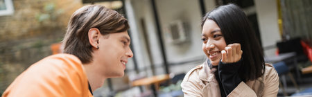 Positive asian woman in trench coat talking to boyfriend in outdoor cafe, banner