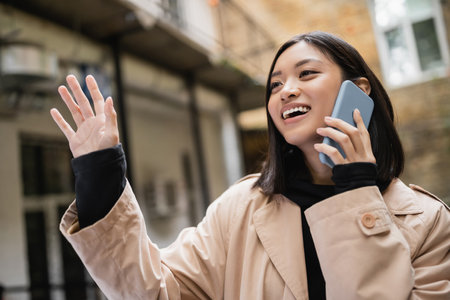 Smiling asian woman talking on smartphone and waving hand outdoorsの写真素材