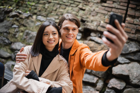 Smiling man hugging asian girlfriend while taking selfie on smartphone on terrace of cafe