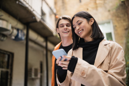 Smiling asian woman in trench coat using smartphone near blurred boyfriend on terrace of cafeの写真素材