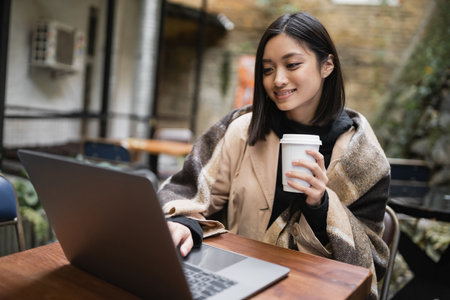Smiling asian woman in blanket holding coffee to go and using laptop on terrace of cafeの写真素材