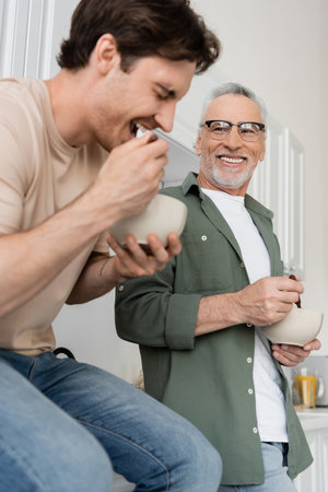happy man in eyeglasses looking at young son having breakfast and laughing on blurred foregroundの写真素材