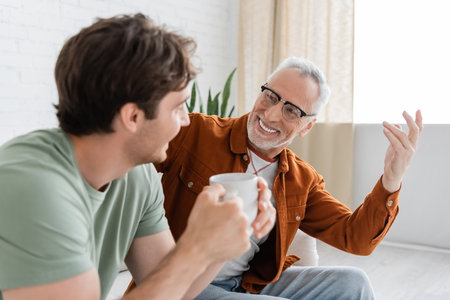 cheerful senior man in eyeglasses gesturing and talking to adult son holding tea cup on blurred...